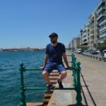 A smiling man in a blue t-shirt, shorts, and cap sits on a green railing along the Thessaloniki waterfront. The blue waters of the Thermaikos Gulf are on one side, with a busy street and apartment buildings on the other.
