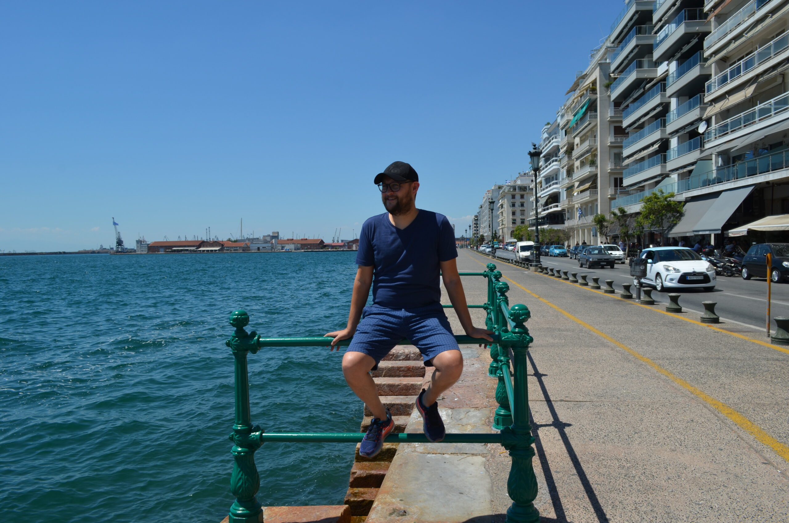A smiling man in a blue t-shirt, shorts, and cap sits on a green railing along the Thessaloniki waterfront. The blue waters of the Thermaikos Gulf are on one side, with a busy street and apartment buildings on the other.