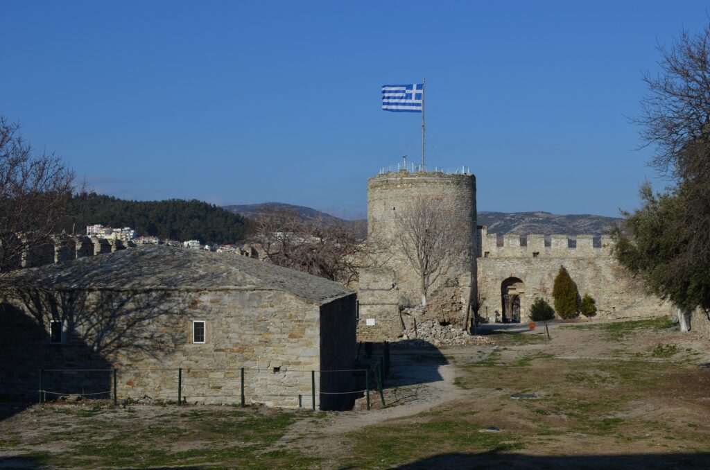 Things to Do in Kavala - A view inside the stone courtyard of the ancient Kavala Castle in Greece, featuring a round tower with a Greek flag, a nearby historic building, and a clear blue sky.