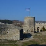 Kavala'da Gezilecek Yerler - Yunanistan'daki Kavala Kalesi'nin iç avlusunun görüntüsü; Yunan bayrağı taşıyan yuvarlak kule, yakındaki tarihi yapı ve berrak mavi gökyüzü.