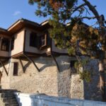 Things to do in Kavala - Exterior view of the historic Ottoman-style House of Muhammad Ali Pasha in Kavala, Greece, featuring stone walls, wooden bay windows (cumba), and a pine tree against a clear sky.