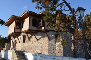Things to do in Kavala - Exterior view of the historic Ottoman-style House of Muhammad Ali Pasha in Kavala, Greece, featuring stone walls, wooden bay windows (cumba), and a pine tree against a clear sky.