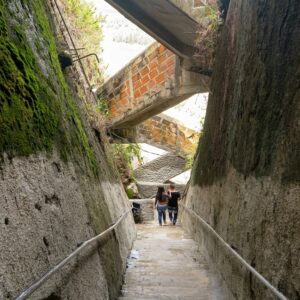 A couple walking down a narrow stone staircase inside a tight vertical fissure of El Peñón de Guatapé rock in Colombia, flanked by high mossy stone walls with crisscrossing brick and concrete staircases above.