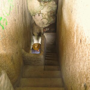 A high-angle view looking down a narrow, steep staircase tunnel enclosed by massive rock walls inside El Peñón de Guatapé, Colombia, showing a tourist wearing a sun hat descending below.