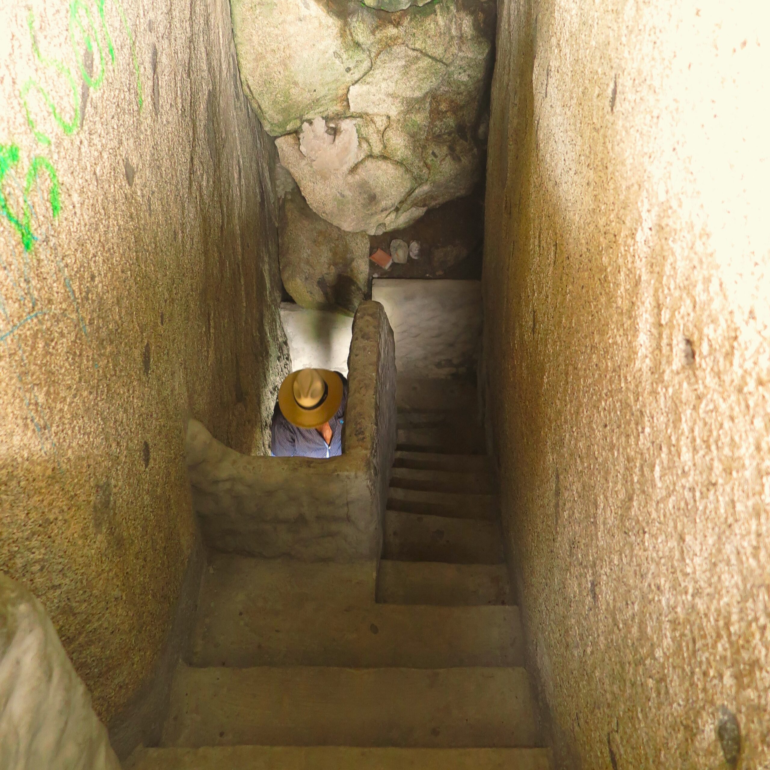 A high-angle view looking down a narrow, steep staircase tunnel enclosed by massive rock walls inside El Peñón de Guatapé, Colombia, showing a tourist wearing a sun hat descending below.