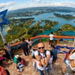 A wide-angle photograph taken from the observation deck on top of the El Peñón de Guatapé monolith in Colombia. In the center, a smiling man is taking a selfie. Behind him is a breathtaking panoramic view of the labyrinthine reservoir filled with many small, lush green islands under a blue sky with white clouds.