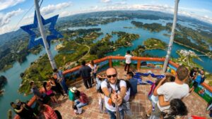 A wide-angle photograph taken from the observation deck on top of the El Peñón de Guatapé monolith in Colombia. In the center, a smiling man is taking a selfie. Behind him is a breathtaking panoramic view of the labyrinthine reservoir filled with many small, lush green islands under a blue sky with white clouds.