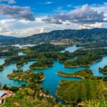 A high-angle, vibrant landscape photograph taken from the top of El Peñón de Guatapé, Colombia. The shot showcases the Guatapé reservoir with turquoise blue waters weaving around lush green islands, peninsulas, and rolling green hills under a cloud-filled sky.