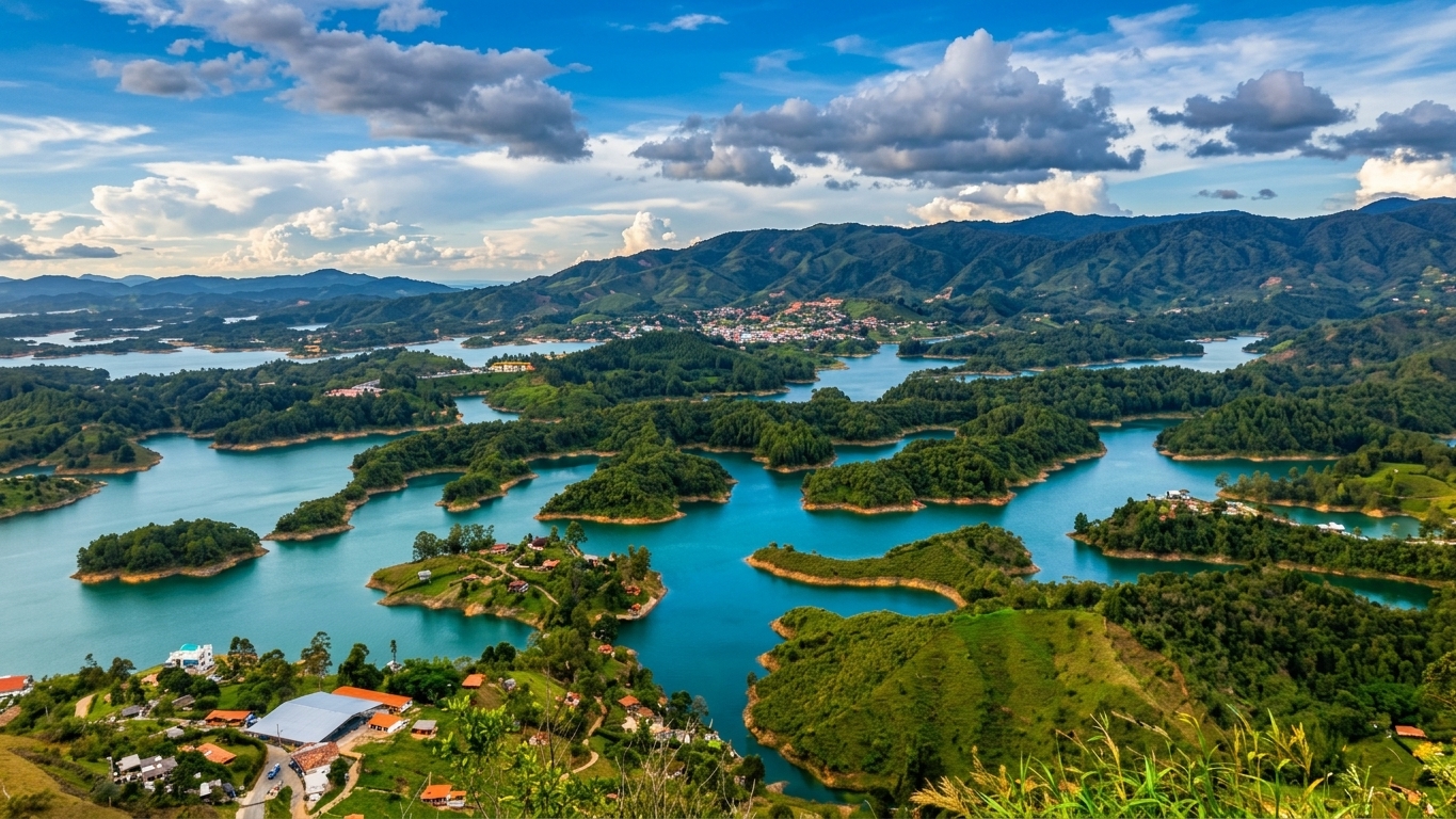 A high-angle, vibrant landscape photograph taken from the top of El Peñón de Guatapé, Colombia. The shot showcases the Guatapé reservoir with turquoise blue waters weaving around lush green islands, peninsulas, and rolling green hills under a cloud-filled sky.