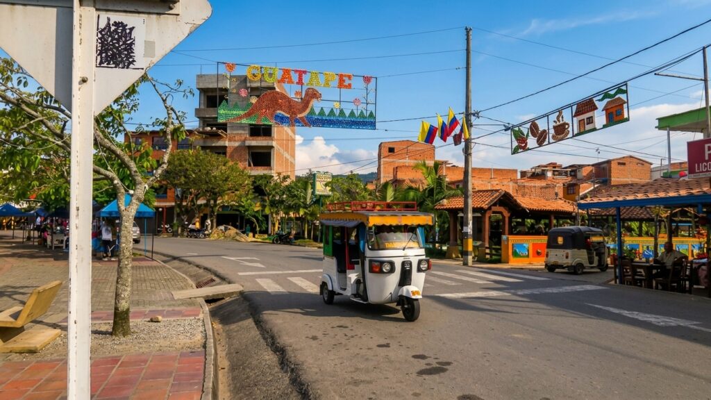 Guatapé Colombia 2 A white and yellow tuk-tuk driving down a paved street in Guatapé, Colombia. Above the street hangs a colorful decorative "GUATAPÉ" welcome sign, Colombian flags, and coffee-themed decorations against a backdrop of local brick architecture.