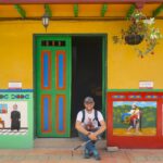 A smiling male tourist sitting in the doorway of a colorful zócalo-decorated house in Guatapé, Colombia.