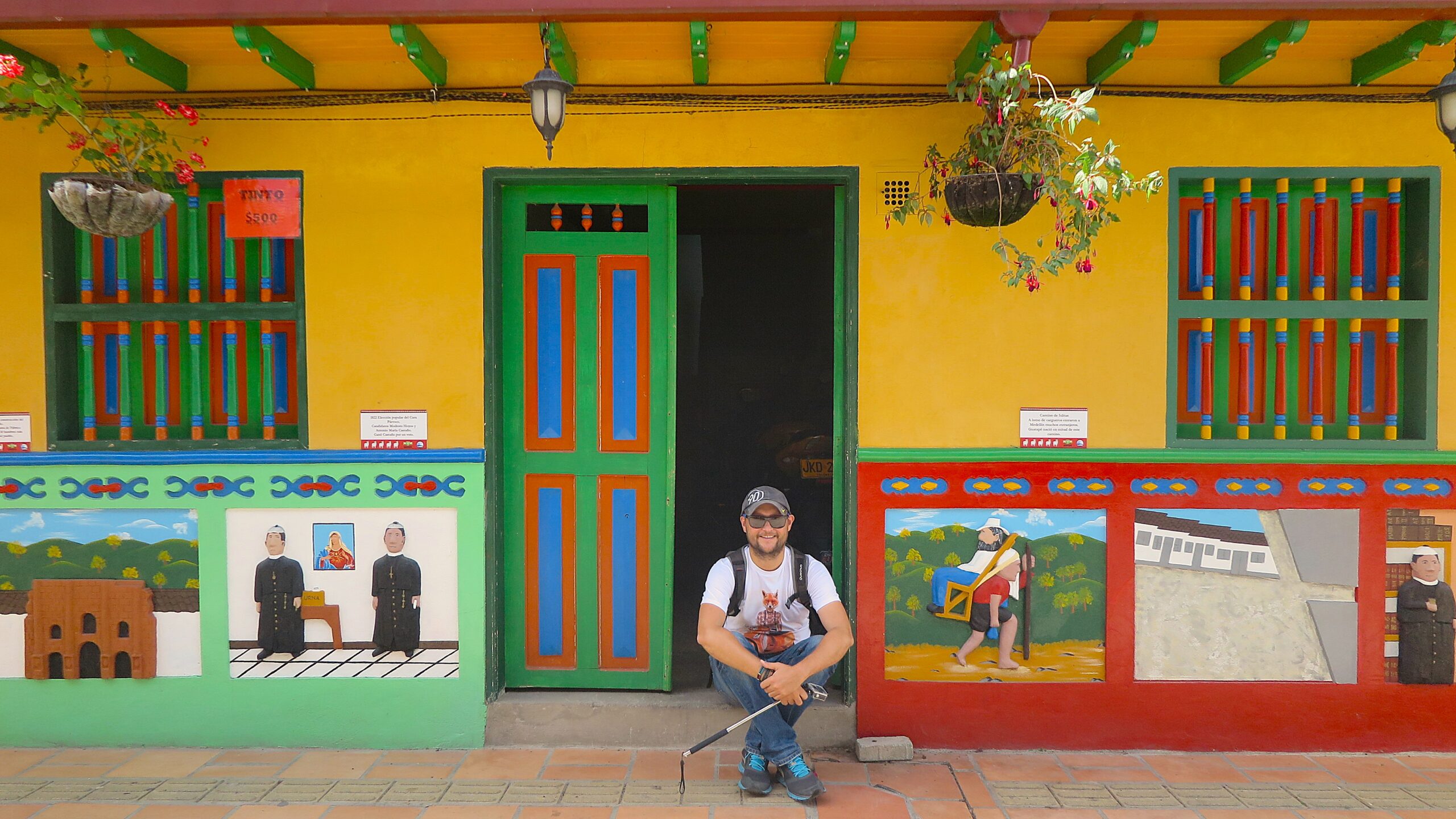A smiling male tourist sitting in the doorway of a colorful zócalo-decorated house in Guatapé, Colombia.
