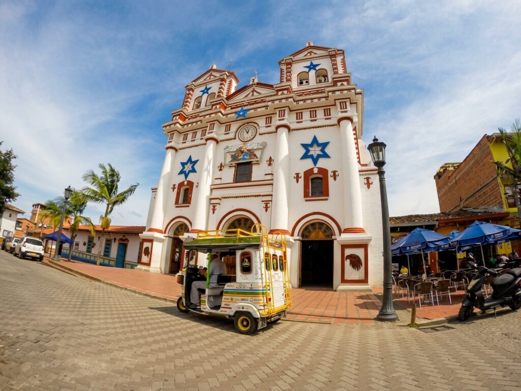 Guatapé Nuestra Señora del Carmen Church A colorful white church in Guatapé, Colombia, featuring red trim and prominent blue stars. A three-wheeled motorbike taxi (tuk-tuk) is parked in the foreground. Palm trees and other colonial buildings are nearby.