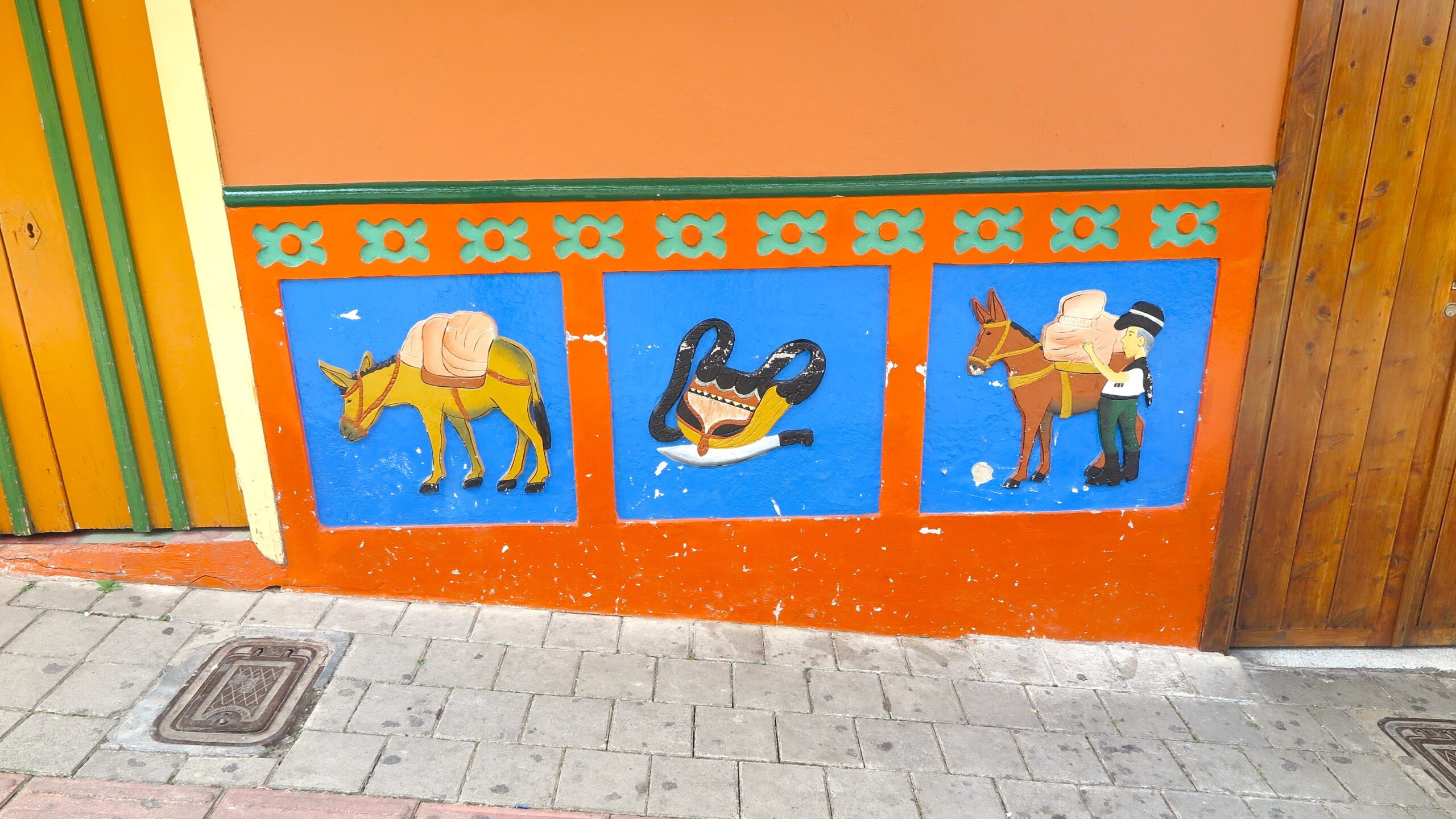 Colorful hand-painted baseboards, known as zócalos, featuring a pack donkey, a traditional bag and machete, and a farmer with a donkey on a bright orange wall in Guatapé, Colombia.