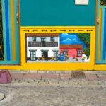 Vibrant teal and yellow building facade in Guatapé, Colombia, featuring a relief zócalo depicting a traditional street scene with colonial houses and playing children.