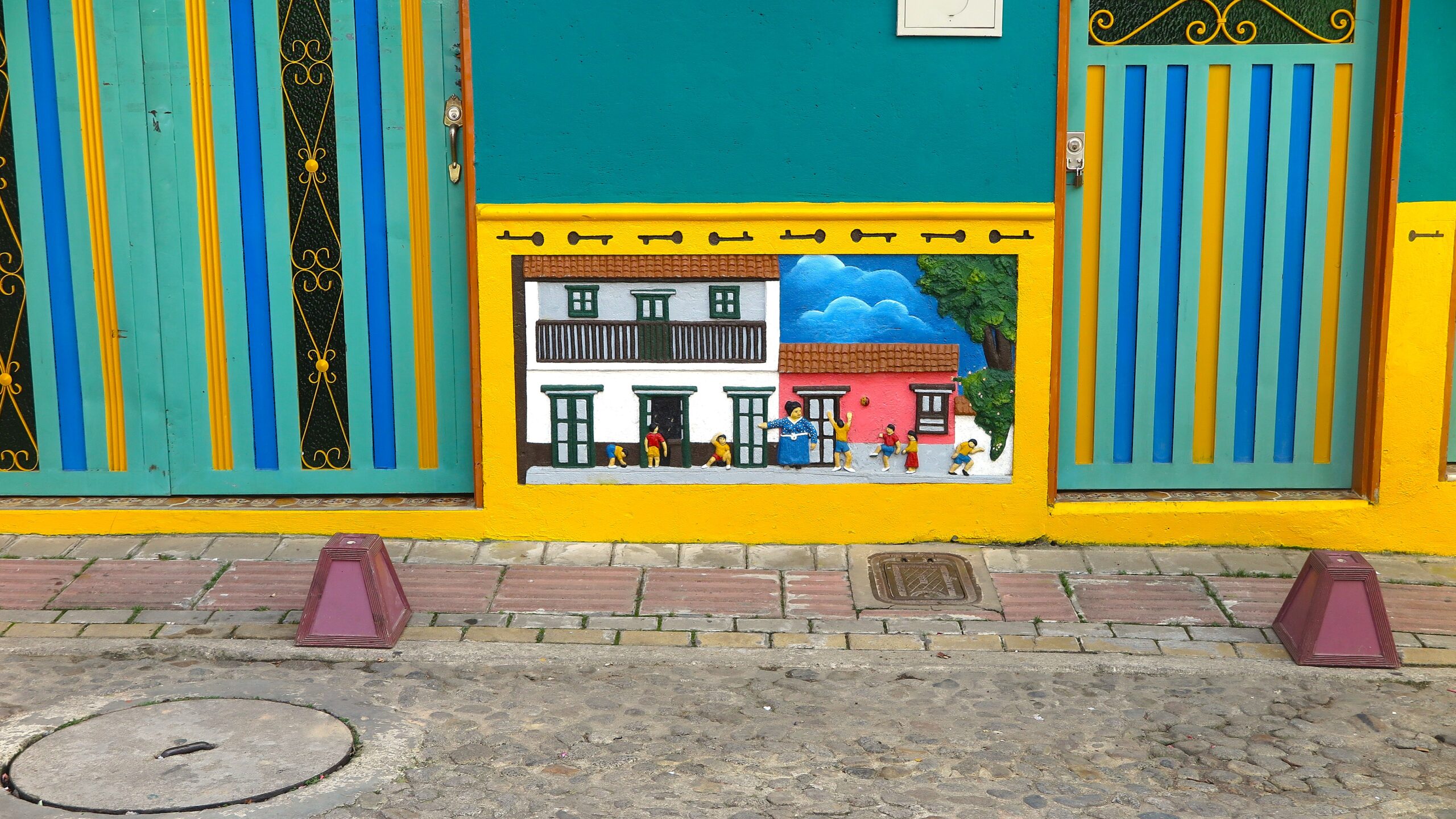 Vibrant teal and yellow building facade in Guatapé, Colombia, featuring a relief zócalo depicting a traditional street scene with colonial houses and playing children.