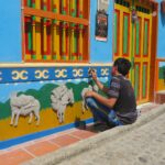 An artisan sitting on a cobblestone street in Guatapé, Colombia, carefully hand-painting relief donkey figures on a bright blue and yellow zócalo panel.