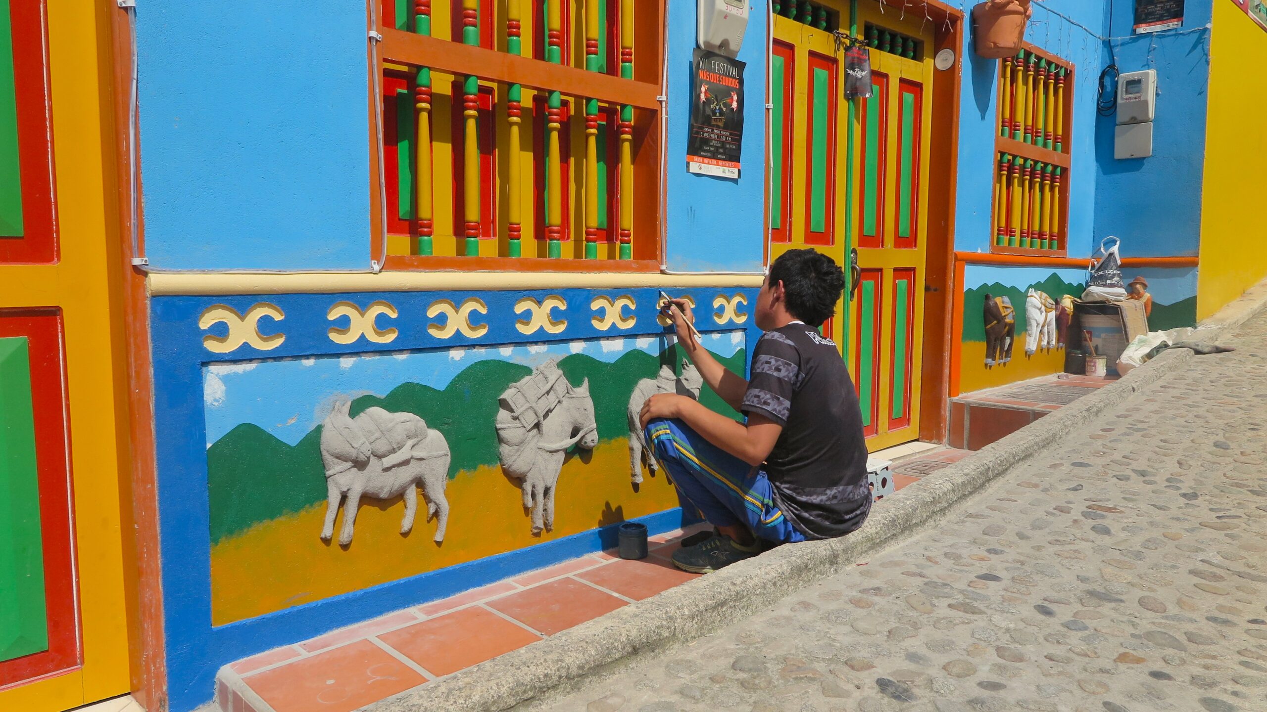 An artisan sitting on a cobblestone street in Guatapé, Colombia, carefully hand-painting relief donkey figures on a bright blue and yellow zócalo panel.