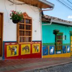 Picturesque cobblestone street in Guatapé, Colombia, lined with brightly painted white, turquoise, and green houses featuring wooden doors and traditional zócalo wall panels depicting donkeys and llama figures.