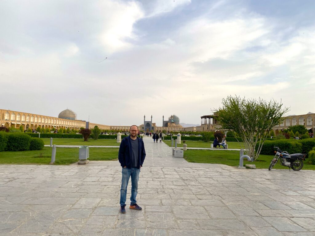 A man wearing glasses and a navy cardigan standing in the historic Naqsh-e Jahan Square in Isfahan, with the Shah Mosque in the background.