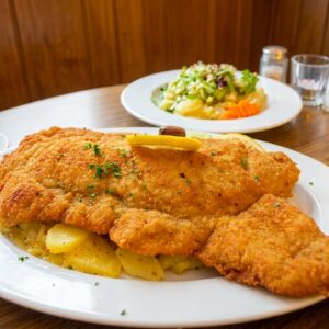 A giant, golden-brown breaded schnitzel served over potato salad with a fresh side salad at Bei Oma Kleinmann, featured in a guide on where to eat in Cologne.