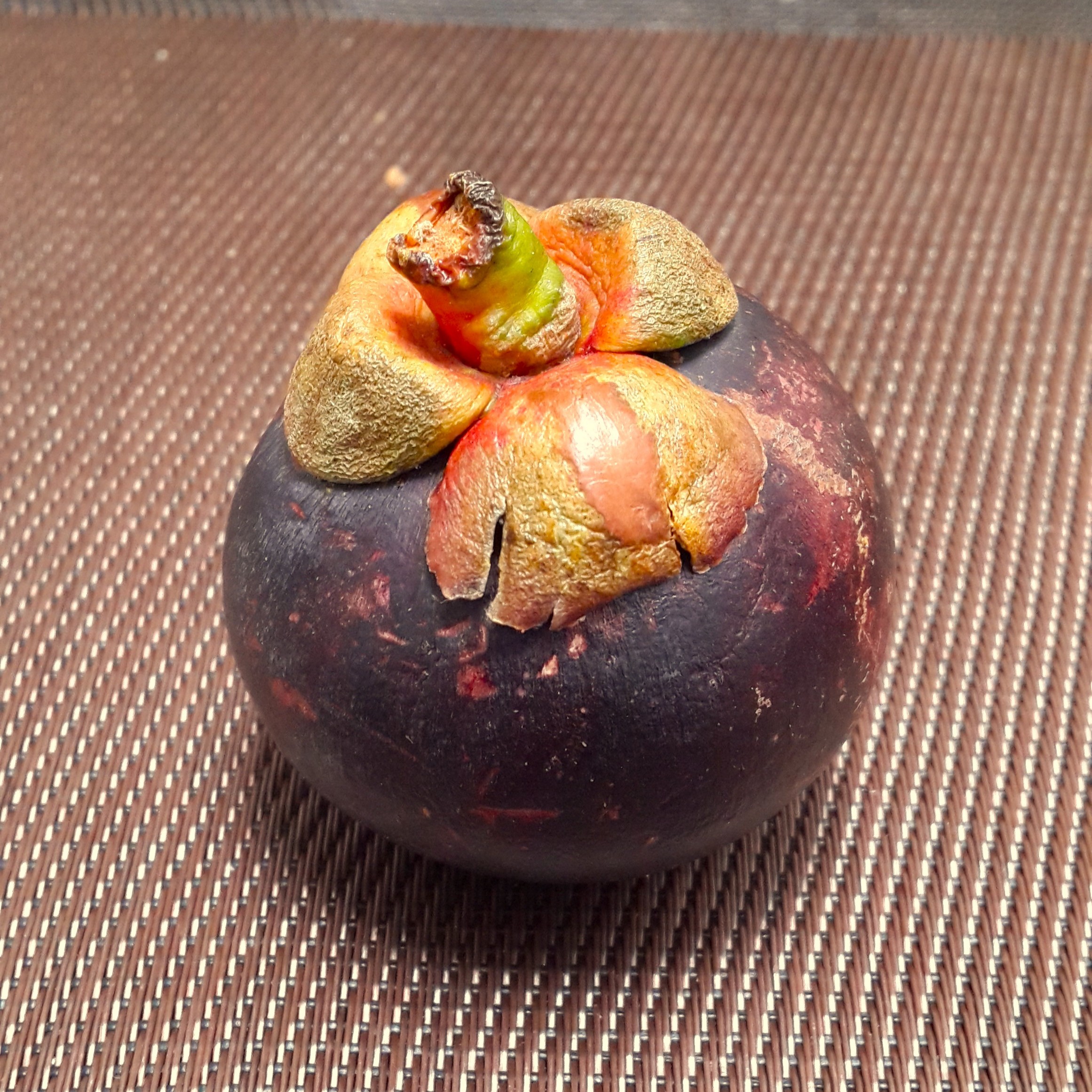 A whole round purple mangosteen fruit from Colombia with its thick crown-like green and brownish calyx resting on a textured woven mat surface.